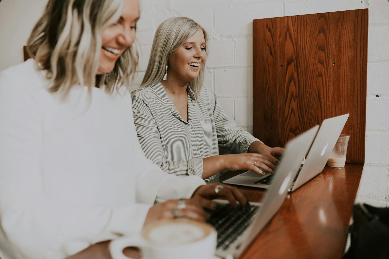Two smiling colleagues work side by side on laptops at a wooden table against a white brick wall.