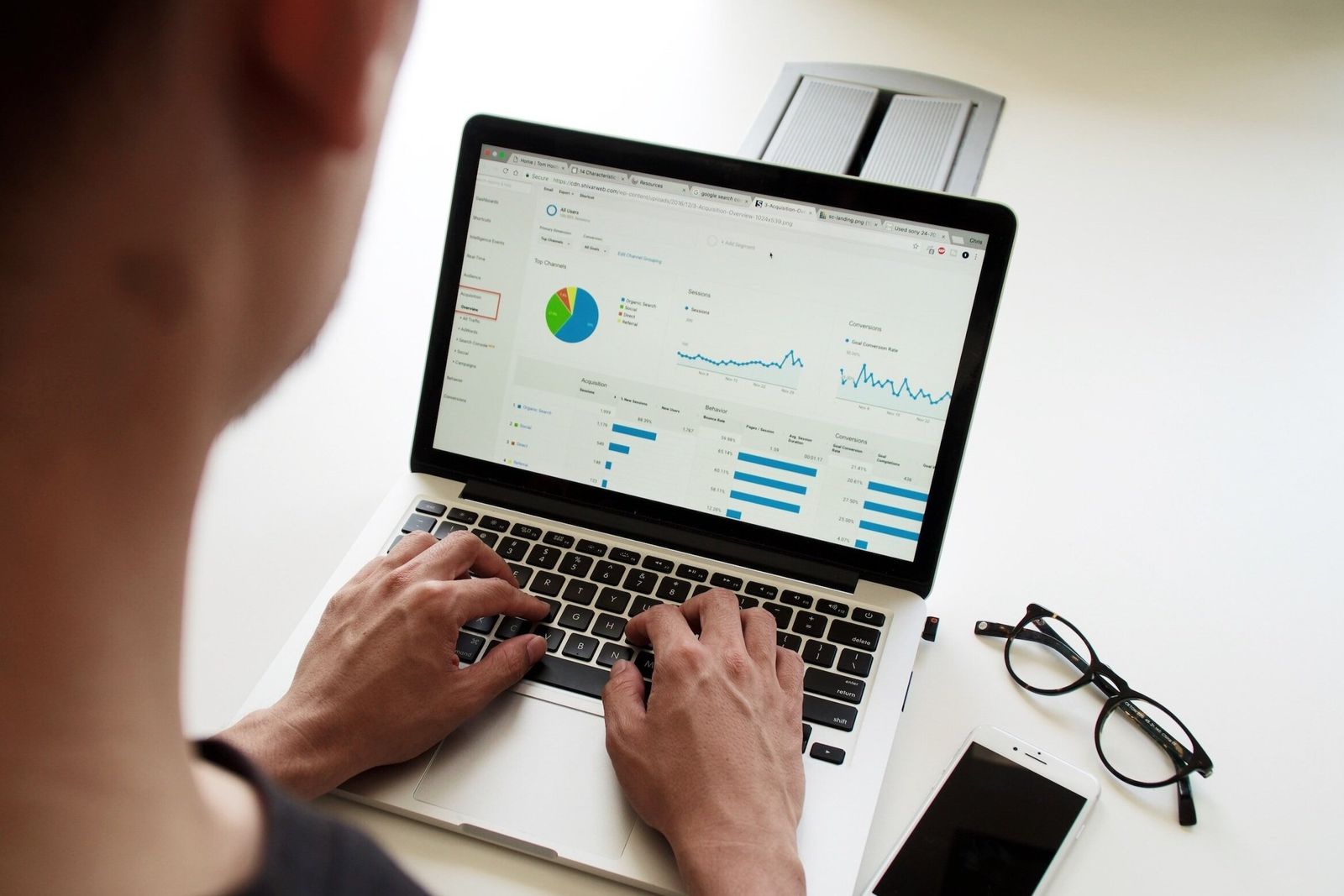 Hands type on a laptop showing charts and dashboards, beside glasses and a phone.