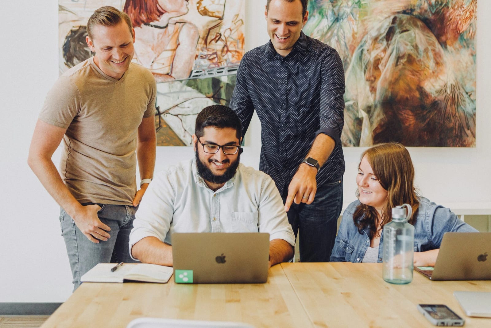 Four colleagues laugh as they gather around a laptop on a wooden table, collaborating on a project.